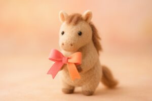 ピンクとオレンジのリボンを持つ手作りフェルト馬の可愛いフィギュア。柔らかなパステル背景で撮影されたブログ用横長の一眼レフ風写真